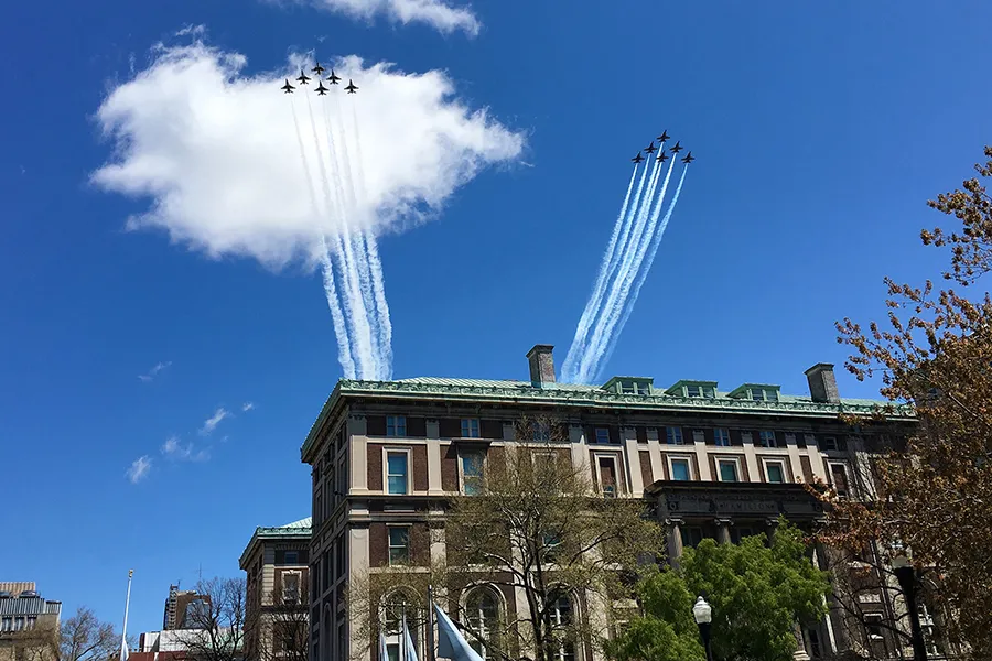 Two teams of six Thunderbird and Blue Angels jet planes fly across a blue sky with clouds over Columbia University's Morningside campus buildings.