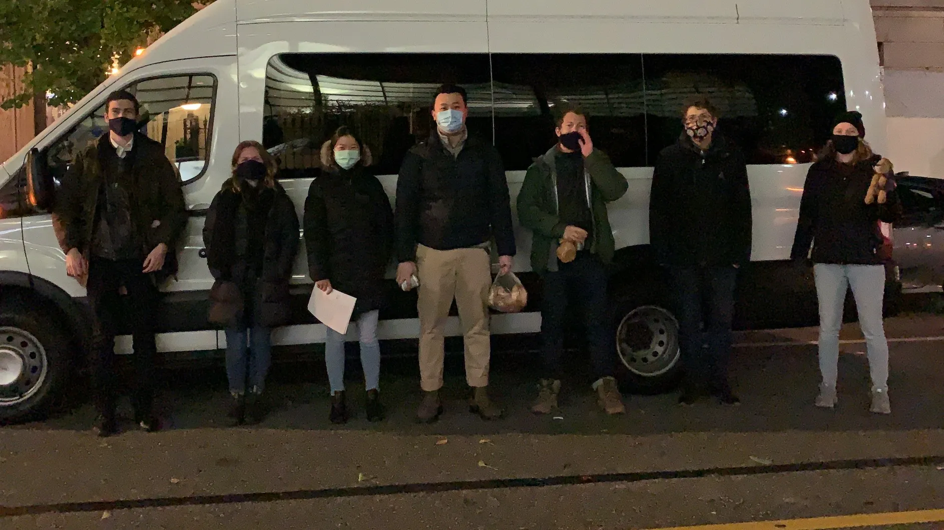 A photo of seven people wearing masks standing in front a white van on the street at night.
