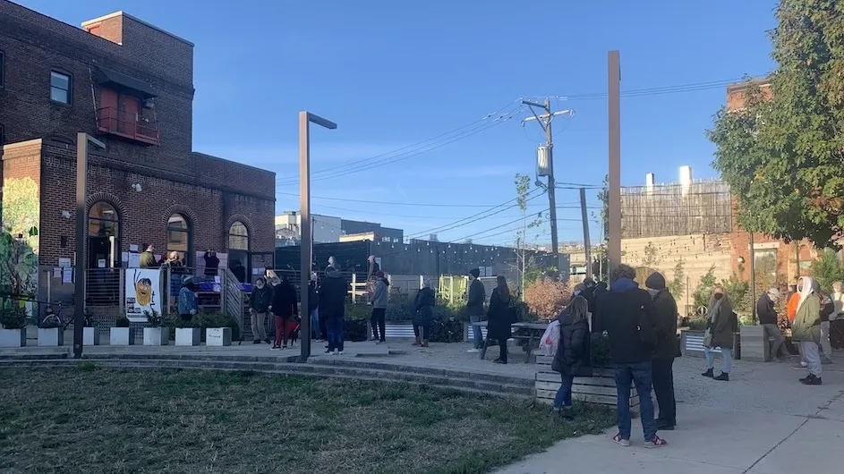 An image of a line of people outside a red brick building on a sunny day.