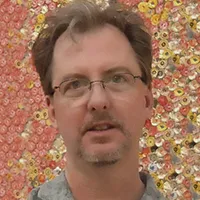 Light-skinned man with medium brown hair and beard, wearing glasses, against a bright pink, yellow, and orange backdrop