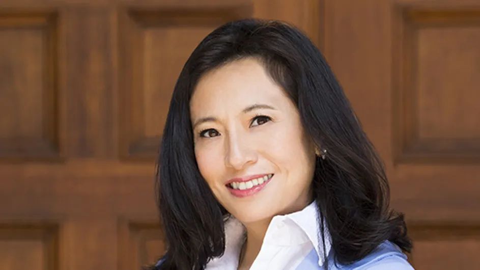 Headshot of a light-skinned woman with long dark hair in a white collared shirt, against a wooden backdrop. 