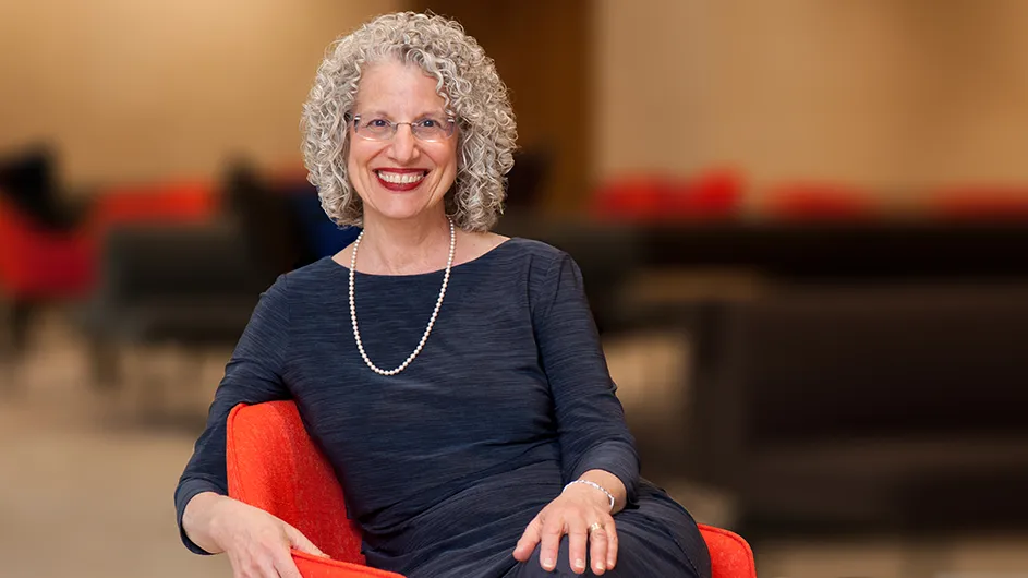 Shuly Rubin Schwartz: a smiling woman wearing glasses, a necklace and a dark top, sitting in a red chair, smiling at the camera.