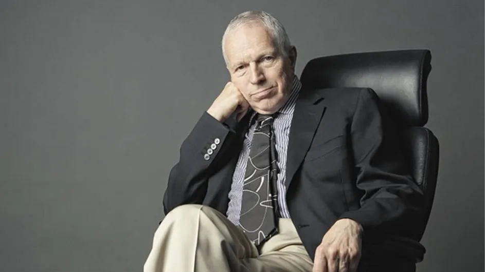 An image of a man wearing a tie and jacket seated in a chair slightly resting his head on his hand.