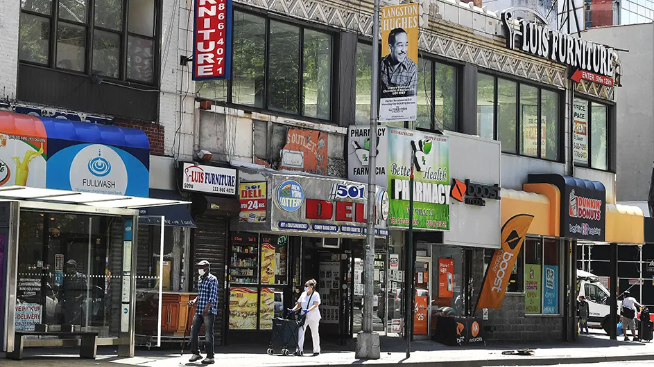 A city street view from summer 2020 in New York City's Harlem neighborhood showing store fronts and people walking and wearing face coverings.