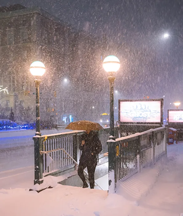 A figure holding an umbrella walks up the stairs from the 116th St. subway entrance onto Columbia's campus. Snow falls all around in the night time.