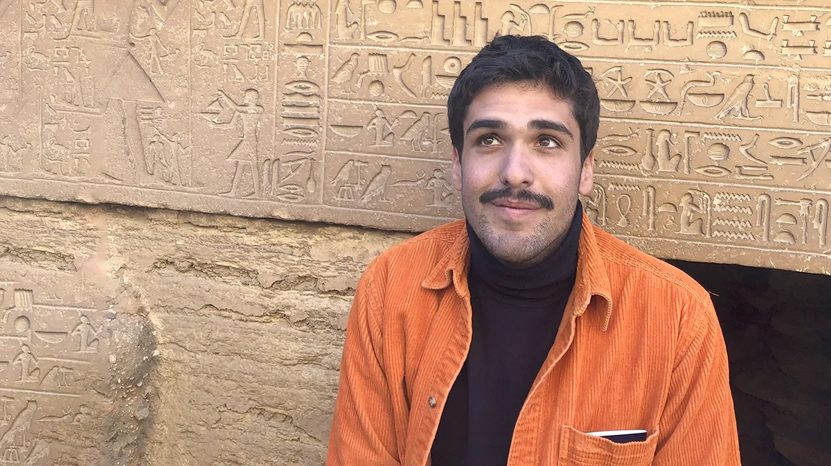 Ibrahim Kombarji: A young man in an orange shirt in front of a wall with carvings.