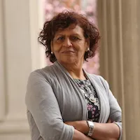 Nabila El-Bassel: A woman with short curly brown hair, standing with her arms crossed in a grey sweater near a columned building.