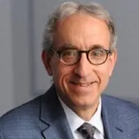 A headshot of a man with grey hair wearing glasses and a grey suit, white shirt, and tie.