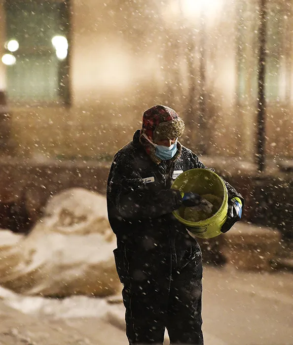 A man in a blue snow suit, blue mask, and fuzzy hat sprinkles snow on Columbia's sidewalks. 