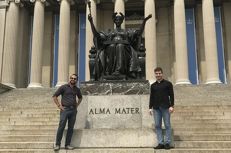 Two men standing on Columbia's Morningside campus, on either side of Alma Mater, a bronze statue of a woman sitting on a throne, with a laurel wreath in her hair and a scepter in her hand.