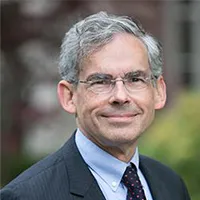 A man in a dark grey suit and grey hair with glasses.