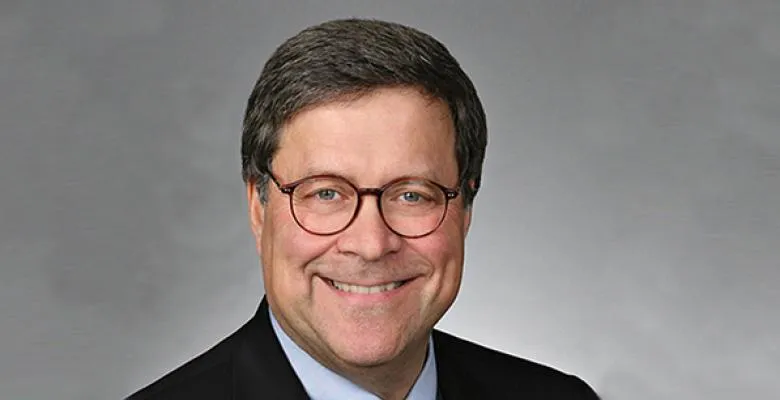 A man, wearing glasses and a dark suit, smiles into the camera against a gray background