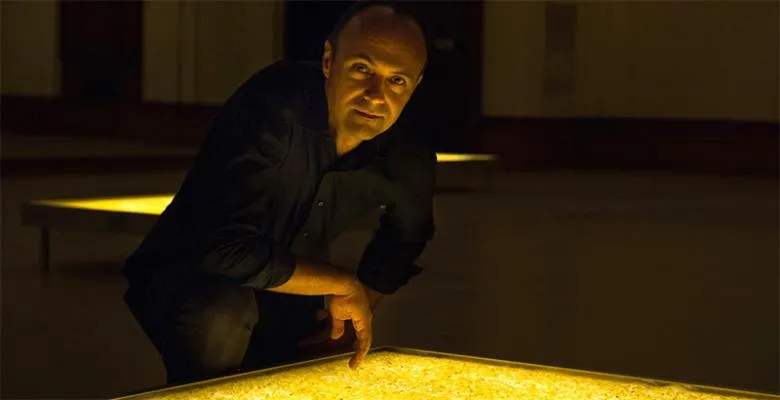 A man wearing a black shirt and black pants leans over a flat table, the surface of which is backlit with amber light