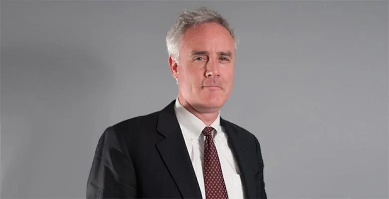 Wearing a dark suit and white shirt, a man stand against a gray background, looking into the camera