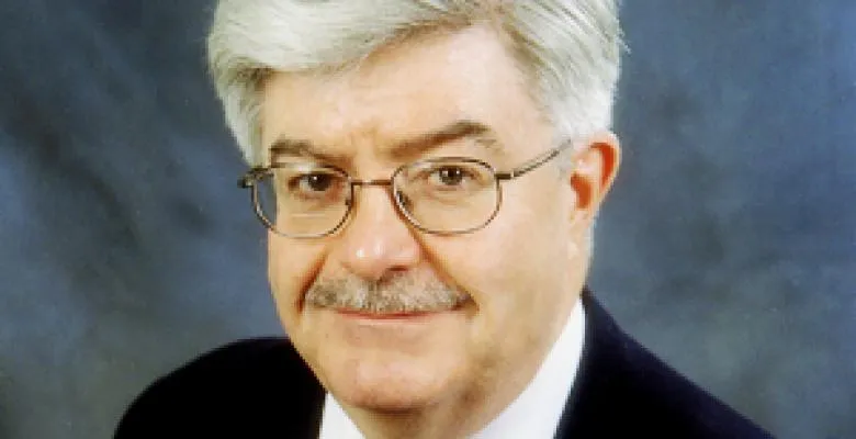 A man with gray hair, mustache and glasses smiles at the camera