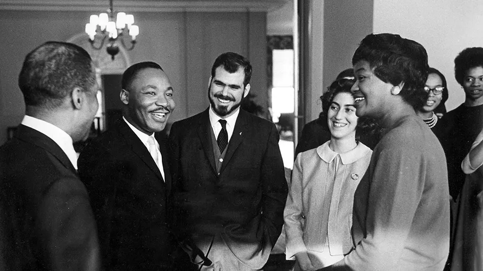 Martin Luther King Jr. in a black suit and white tie talks to a group of students at a Columbia University hall.