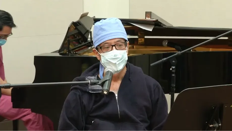 A surgeon with a bonnet and face mask sits at a microphone to sing and has a piano in the background.