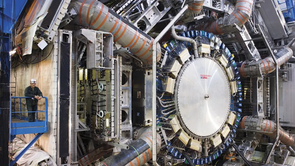 Man on blue platform with hard hat stares out at huge machine - silver cogs like in a watch tinged in blue, red and yello