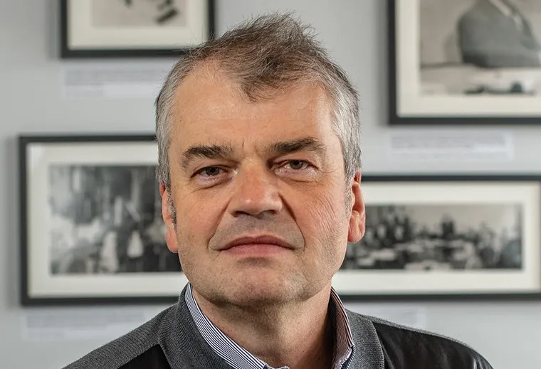 Dmitri Basov: a man with grey hair and fair skin, staring into the camera. Framed black and white pictures are on the wall behind him. 
