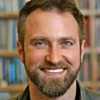Headshot of Caucasian man with short brown hair and beard smiling with bookcases behind him