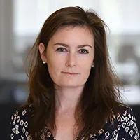 woman in headshot with long brown hair, green eyes and pearl earrings wearing blue dress with diamond white print