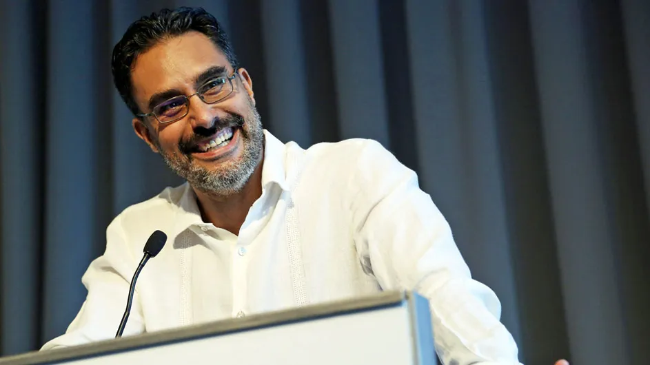 smiling man with glasses, white shirt and brown hair at microphone in front of podium.