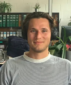 Young man with brown hair wearing a striped shirt and head phones with book case in background