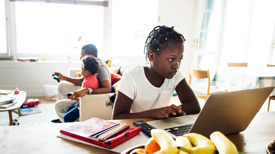 African American middle school aged girl with tight braids and white shirt on a laptop at a desk with father and younger brother (orange shirt) watching TV in background