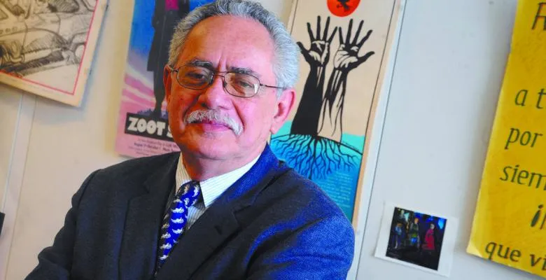 Rodolfo dela Garza looks straight into camera wearning oval shaped eyeglasses short salt and pepper and moustache.  Sporting a blue blazier with a white collared shirt and blue and white necktie standing in front of various pieces of artwork