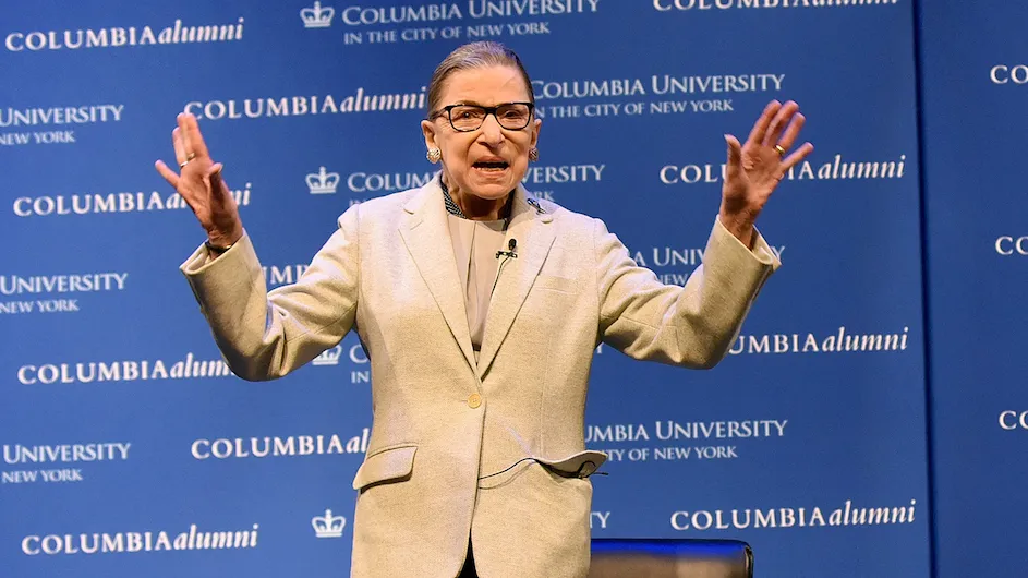 Justice Ruth Bader Ginsburg stands with her arms raised in front of a Columbia University and Columbia Alumni banner
