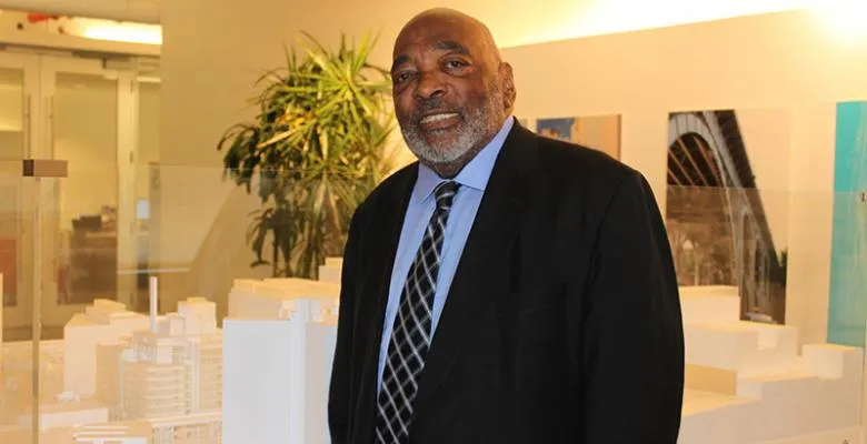 Wilmouth Elmes dressed in dark suit with light blue shirt and dark tie in the lobby of Manhattanville location with model of architectural layout behind him