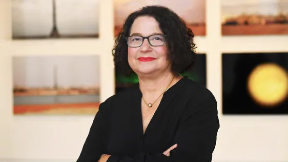 This is an image of a woman with short curly dark hair and glasses in front of paintings.