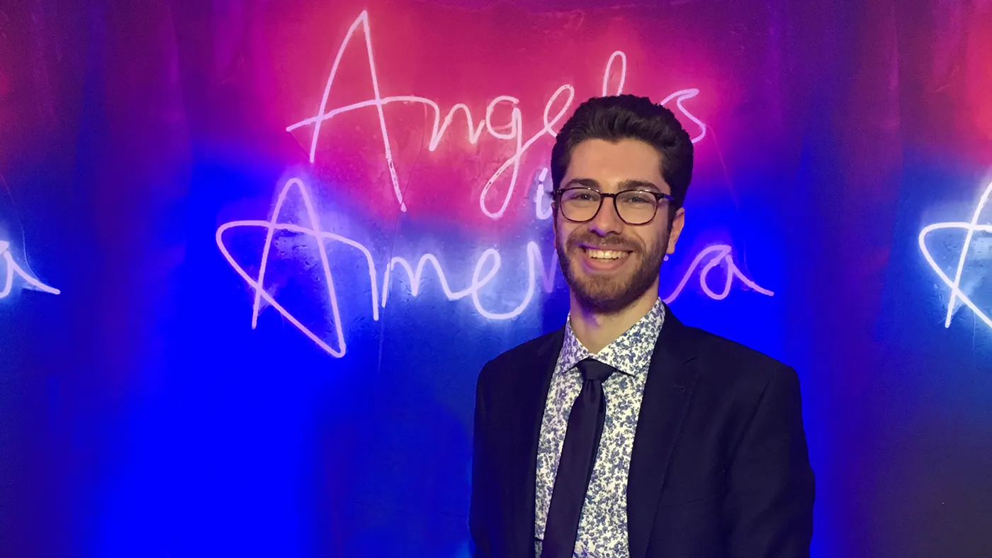 David Treatman: a bespectacled man with dark hair, in jacket, tie and floral shirt, standing in front of a pink and blue backdrop with the words "Angels in America" in cursive across the neon background.
