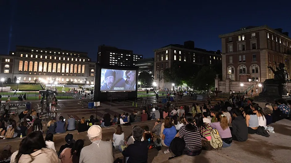 Dozens of students sit on Low steps and watch an outdoor film screening at night with Butler Library in the distance at Columbia University.