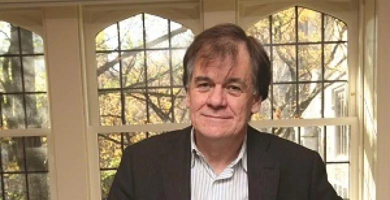 Timothy Mitchell stares into camera lens wearing a dark blazer striped white shirt in front of a country styled window