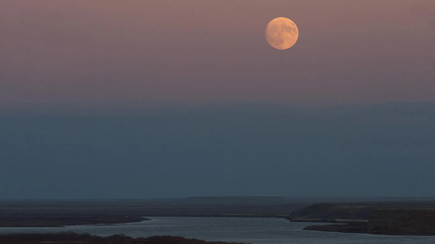 A full pinkish colored moon against a blue and pink sky with barren land and water inlet below.
