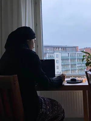 A woman in a dark outfit works at a desk in front of a window through which you can see sky and a building.