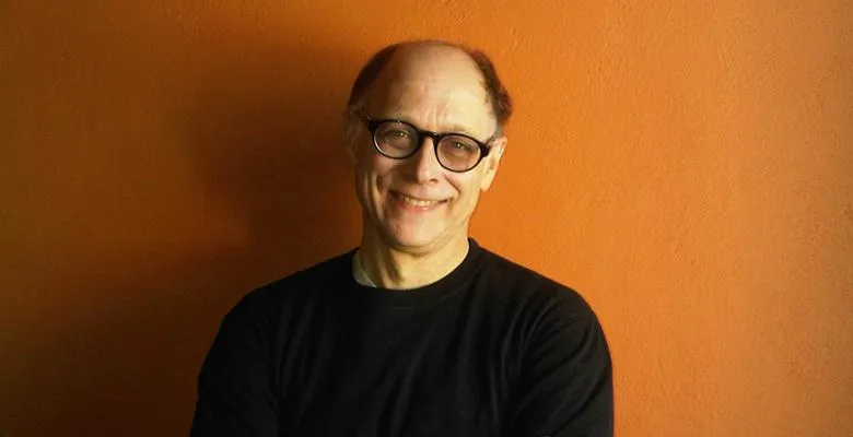 Owen Lewis wearing dark top and dark rimmed eyeglasses while standing against an orange rust like colored wall